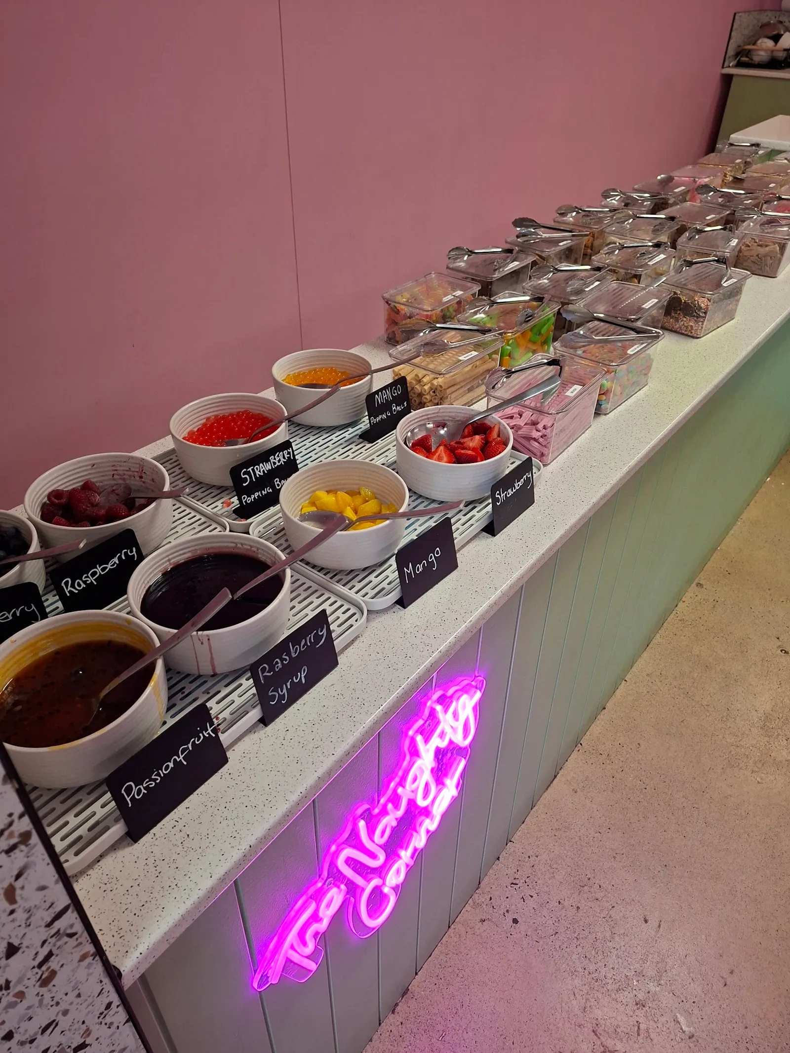 Froyo Heaven toppings bar with labelled fruit and sauce bowls beneath a pink neon sign