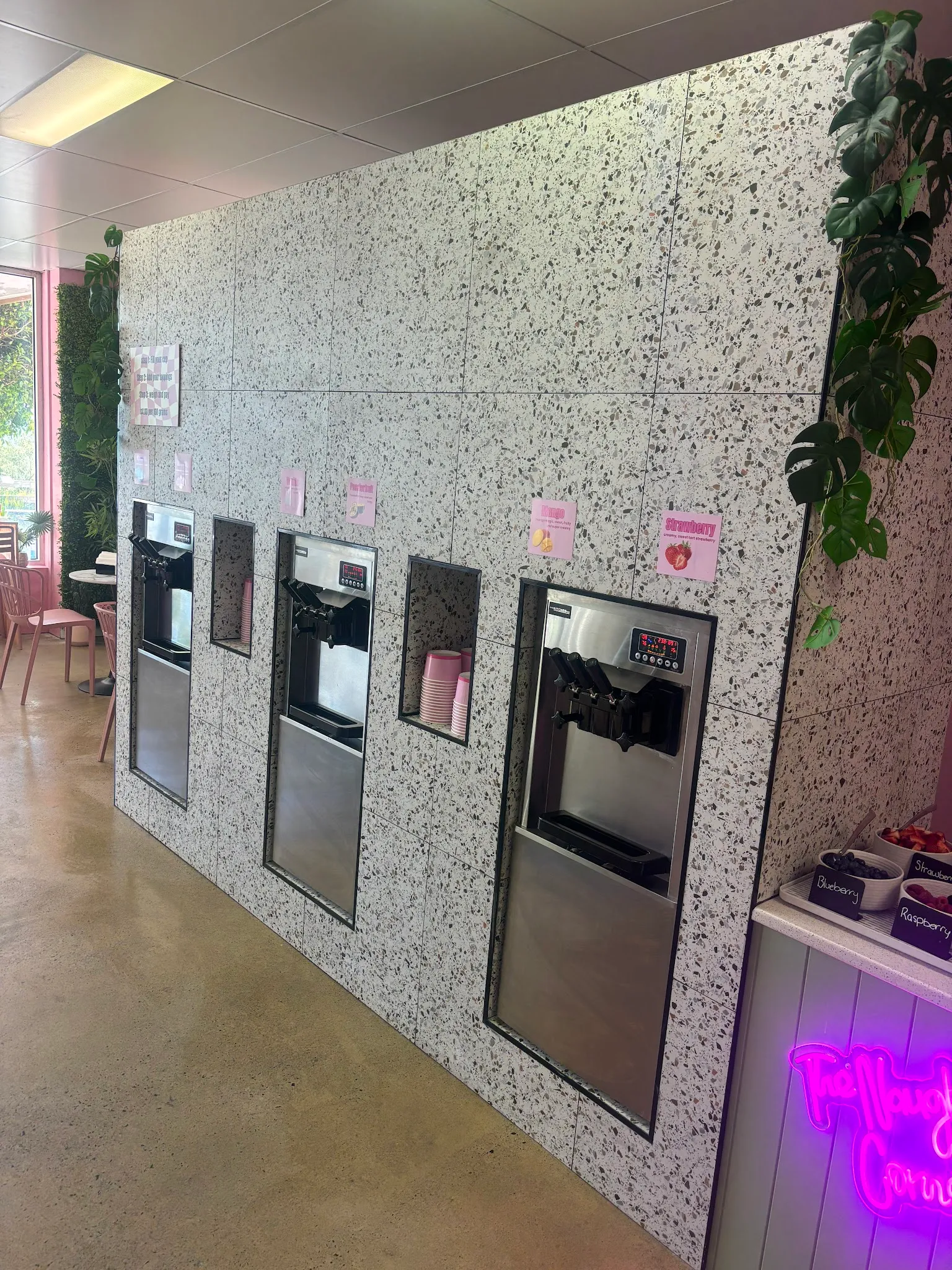 Self-serve yogurt machines set into a terrazzo wall with a pink neon sign below